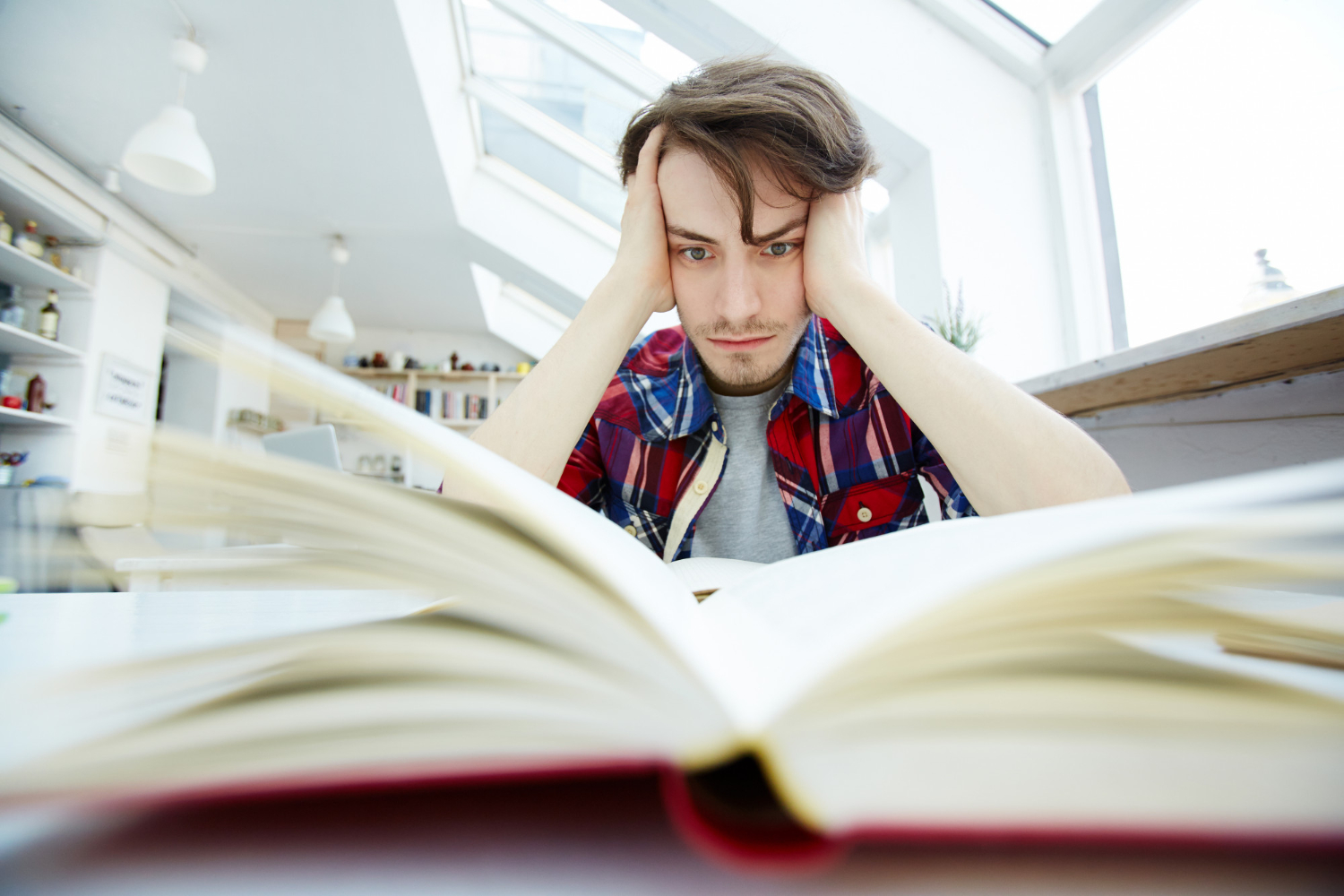 A frustrated man in a plaid shirt stares at an open book in a bright study space.