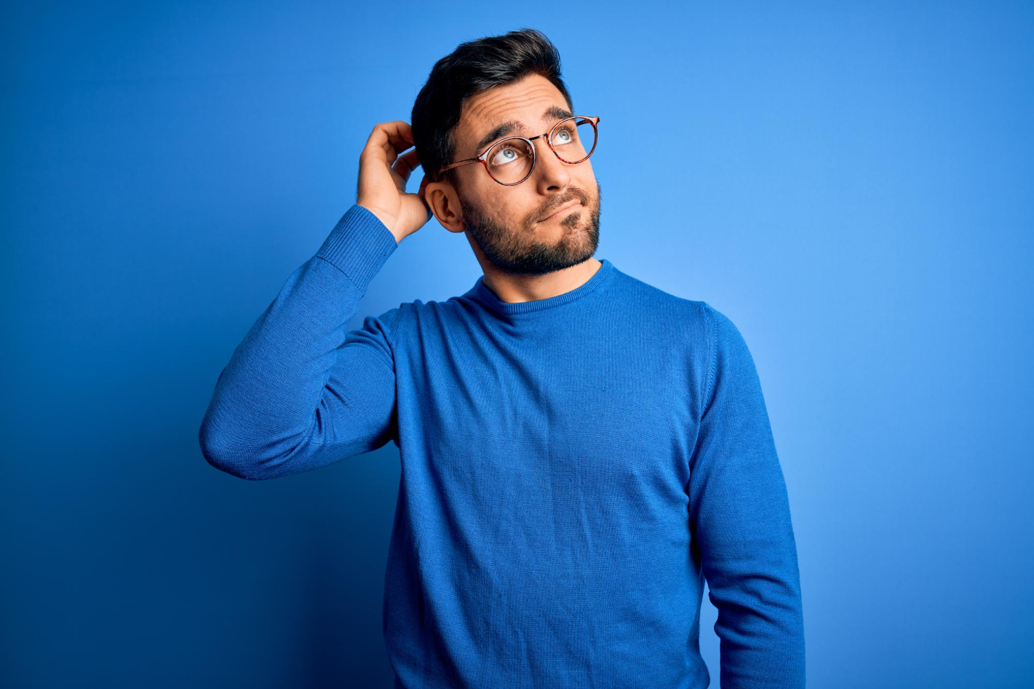 Young handsome man with beard wearing casual sweater and glasses over blue background in thinking