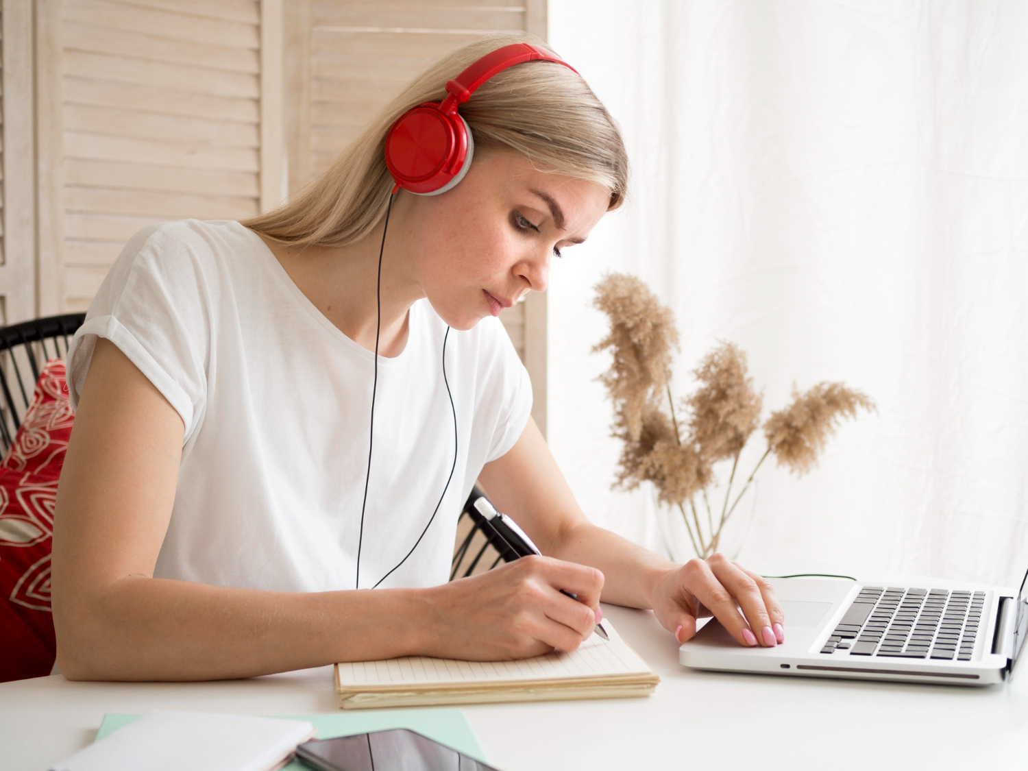 Woman wearing red headphones, writing in a notebook while using a laptop at a desk