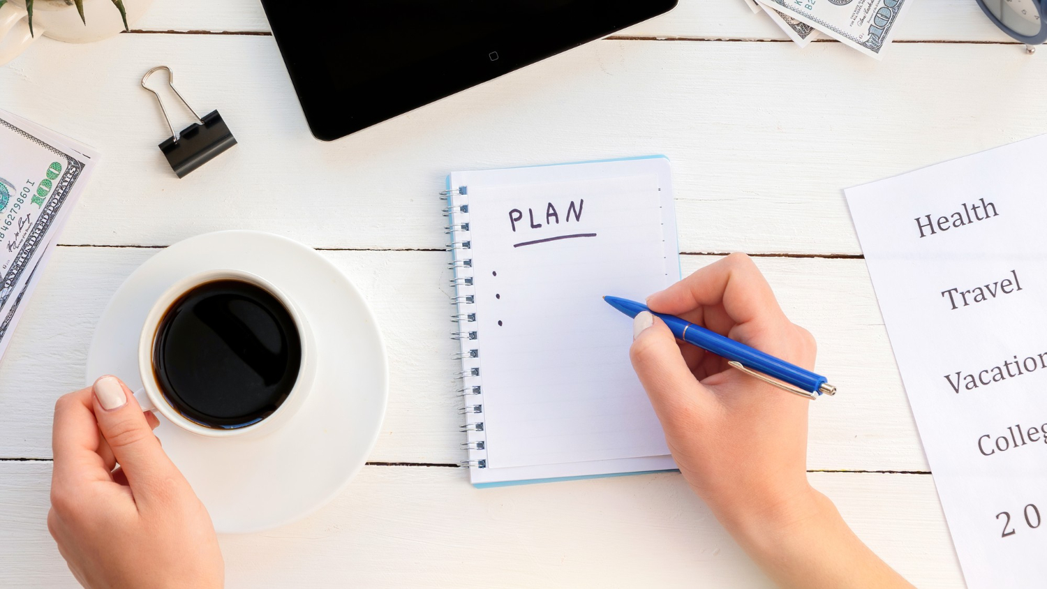 Person writing "PLAN" in a notebook with a pen, next to a cup of coffee, money, and planning documents on a desk.